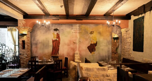 Interior de restaurante con paredes de ladrillo visto, vigas de madera y un mural que representa a dos figuras. Las mesas están dispuestas con copas de vino y cubiertos.