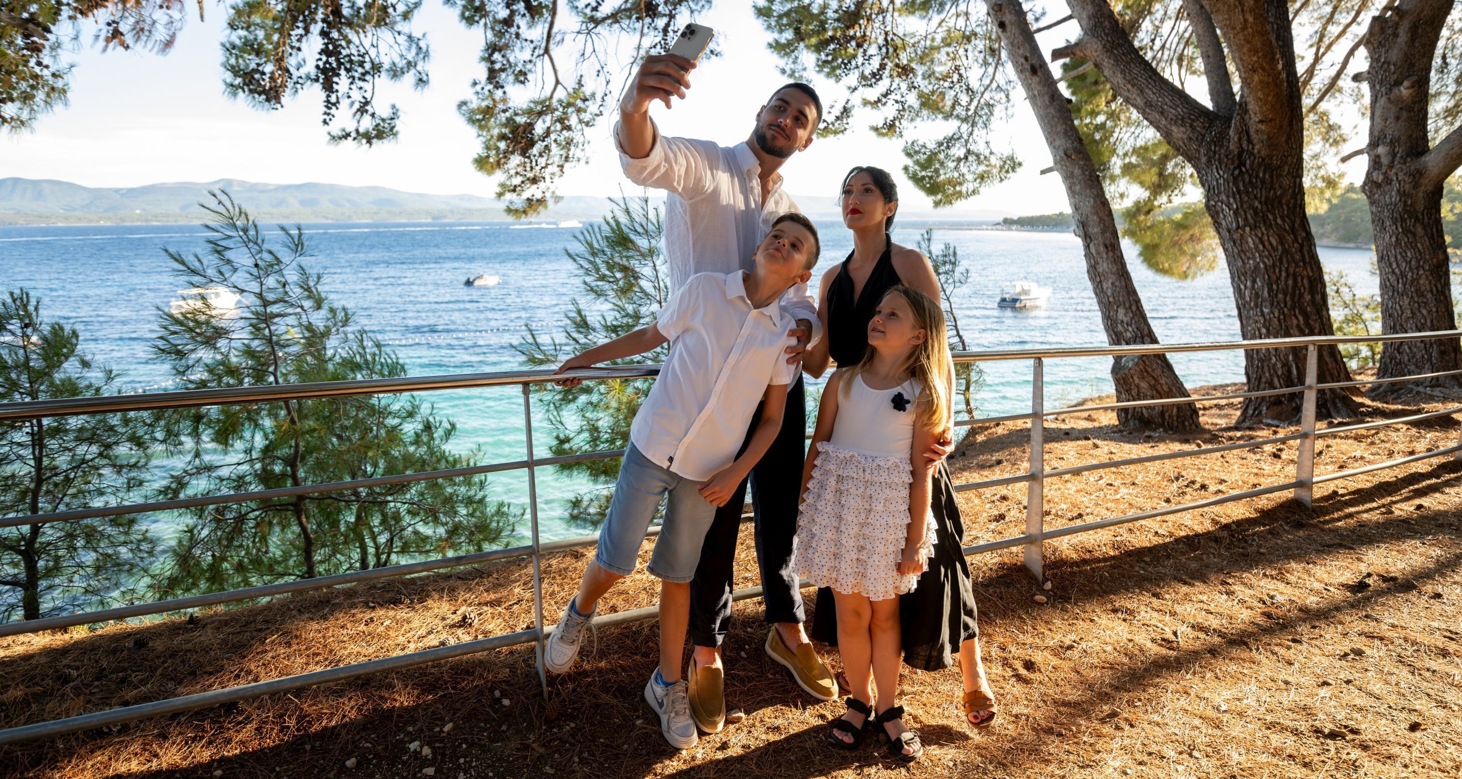 A family taking a selfie by a railing overlooking a body of water with trees and distant mountains.