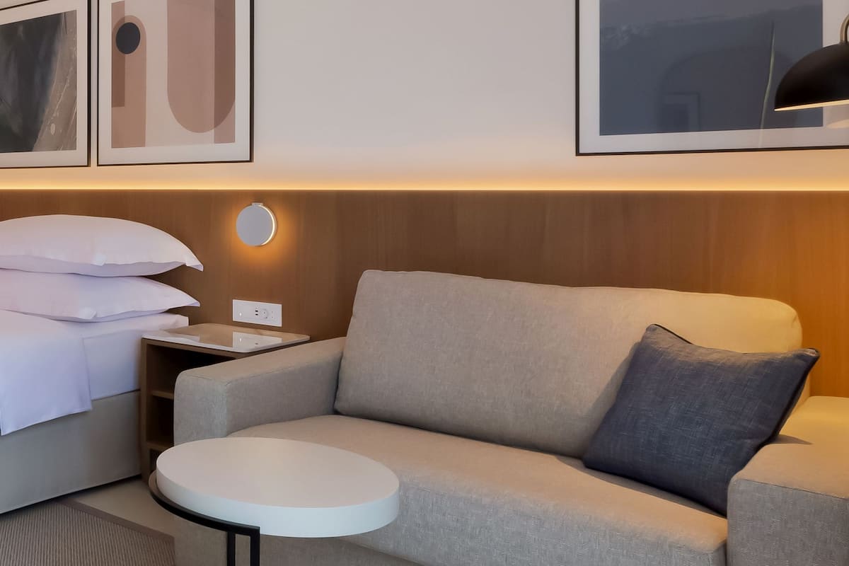 Une chambre d’hôtel avec un lit, un canapé, une petite table et une table de chevet. Des tableaux encadrés ornent le mur et une lampe se trouve sur la table de chevet.