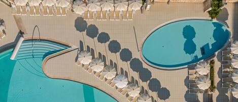 An aerial view of a swimming pool area with multiple sun loungers and umbrellas, a curved pool edge, and a stone-tiled deck.