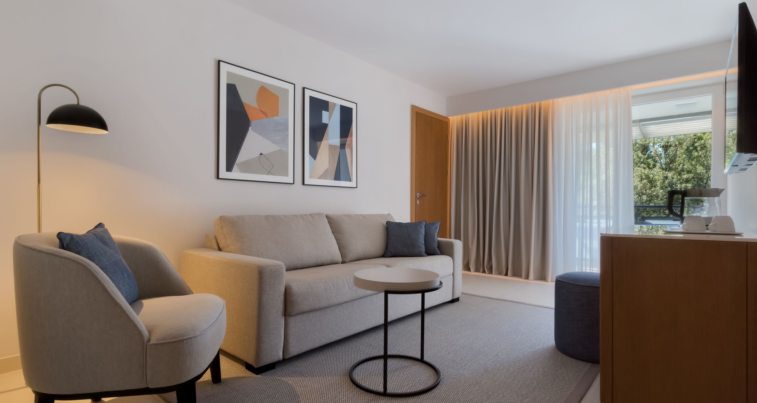 A modern living room with a sofa, armchair, and round coffee table. There are abstract paintings on the wall, a floor lamp, and a view through large windows with curtains.