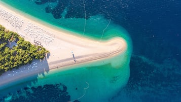 Vista aérea de una playa con costa curva, aguas turquesas cristalinas y densa vegetación verde a la izquierda.
