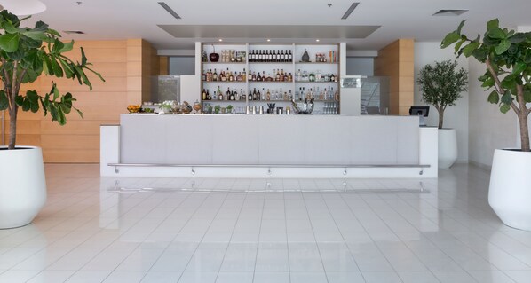 A modern reception area with a white reception desk, a wall of neatly organized liquor bottles, and large potted plants. - Bluesun Hotel Elaphusa (Bol)