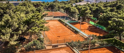 Una cancha de tenis rodeada de árboles y un edificio, con vista al mar y a las montañas al fondo.