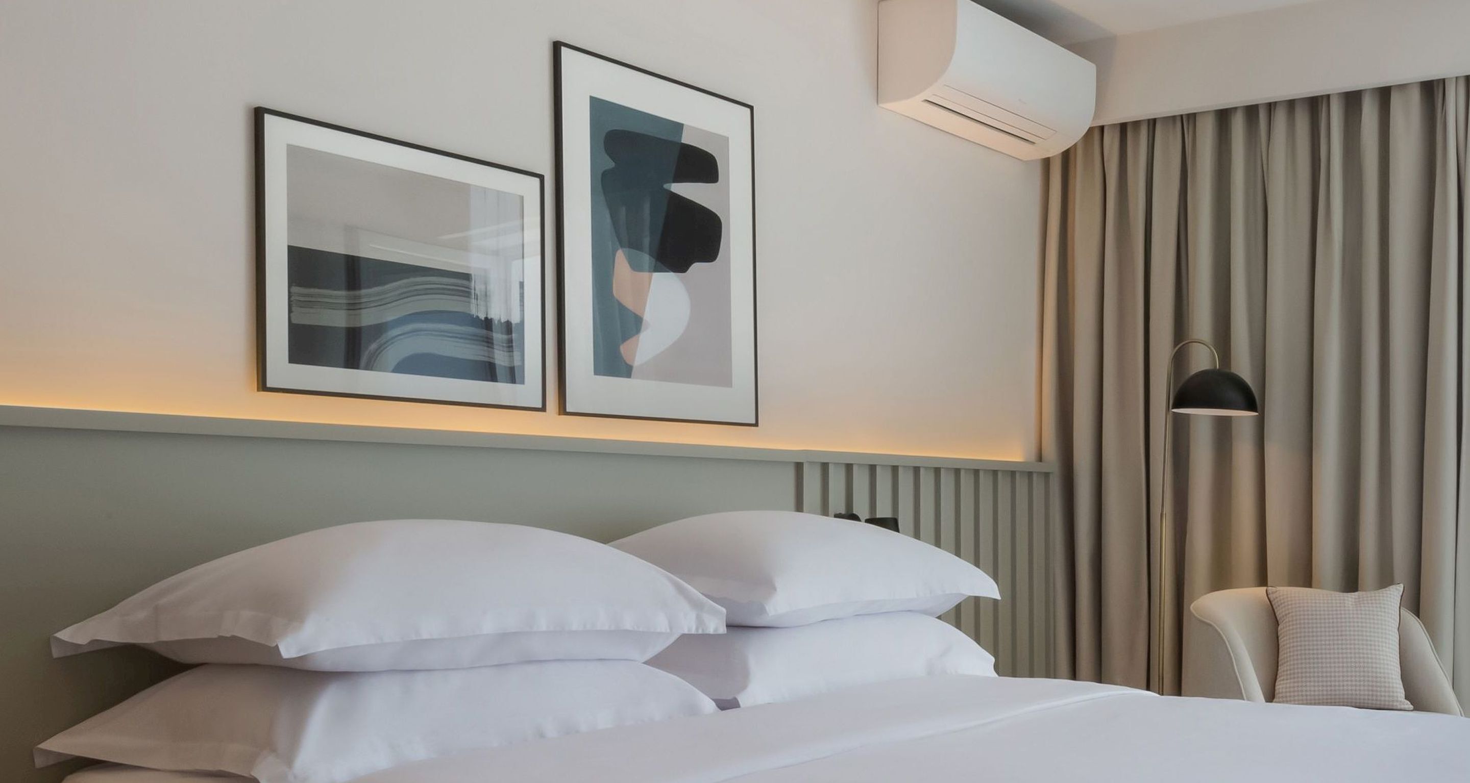 A hotel room with a neatly made bed, two framed artworks on the wall, an air conditioning unit, and a window with curtains.