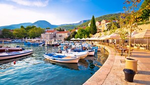 A waterfront marina with boats docked, a stone quay with outdoor seating, and buildings in the background. - Bluesun Hotel Borak (Bol)