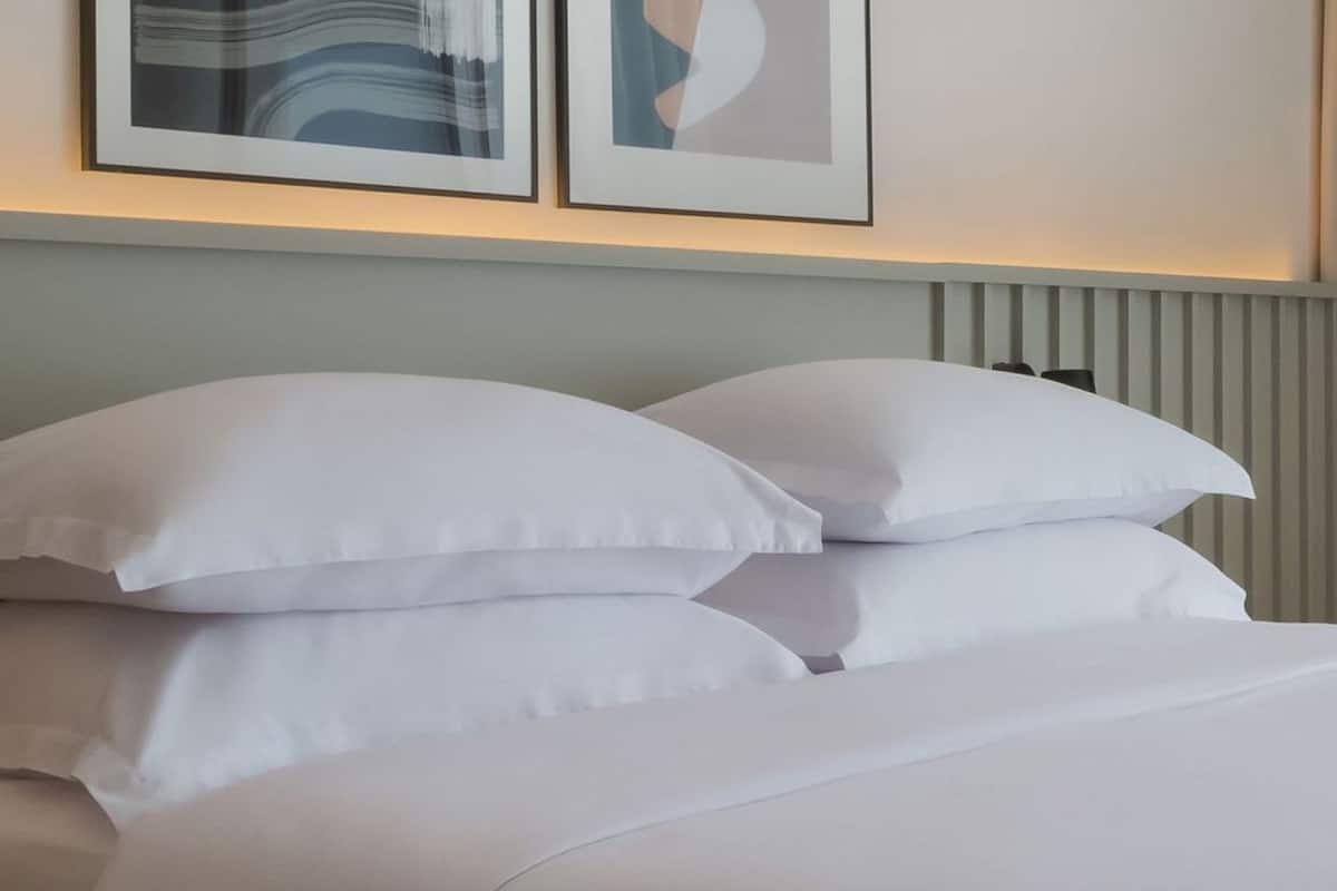 Ein ordentlich bezogenes Bett mit weißer Bettwäsche und Kissen, ein heller Kopfteil und zwei gerahmte Kunstwerke darüber.