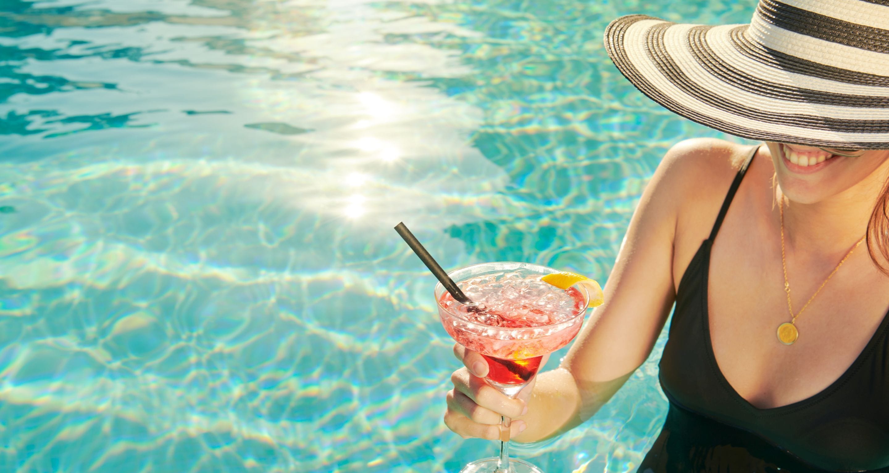 A person holding a cocktail with a straw, wearing a striped hat, and standing by a clear pool.