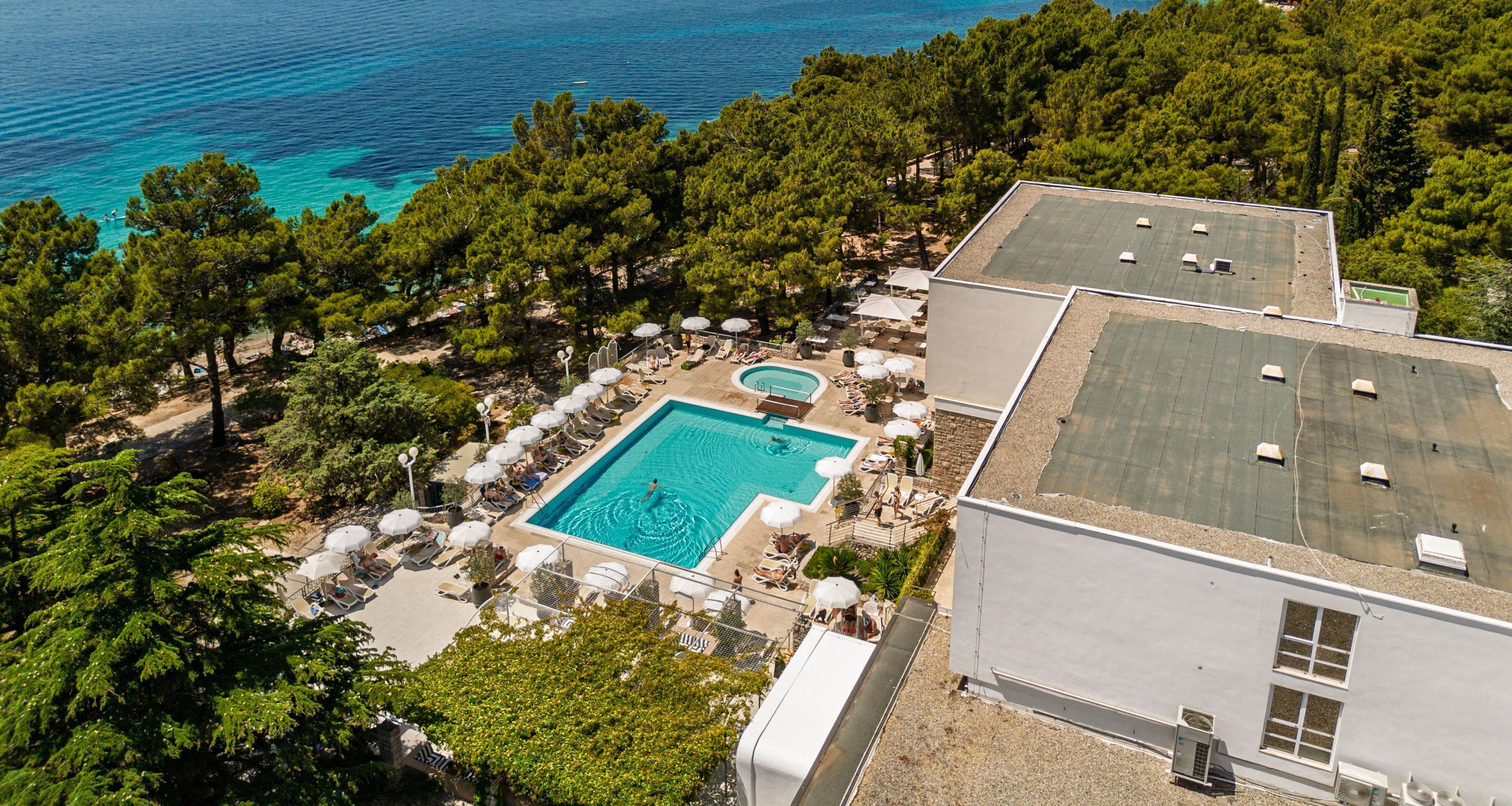 Eine Vogelperspektive zeigt ein Resort mit einem Swimmingpool, umgeben von Liegestühlen und Sonnenschirmen, einen Tennisplatz sowie Blick aufs Meer.