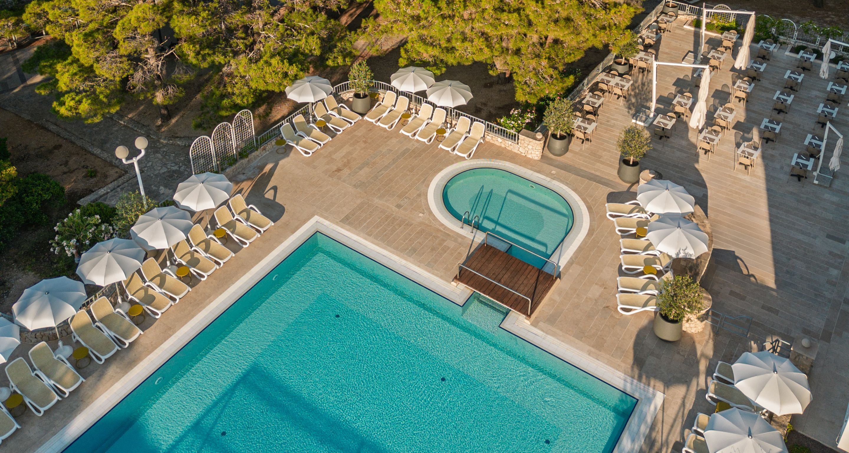 An aerial view of a resort with a central swimming pool, sun loungers, and umbrellas.