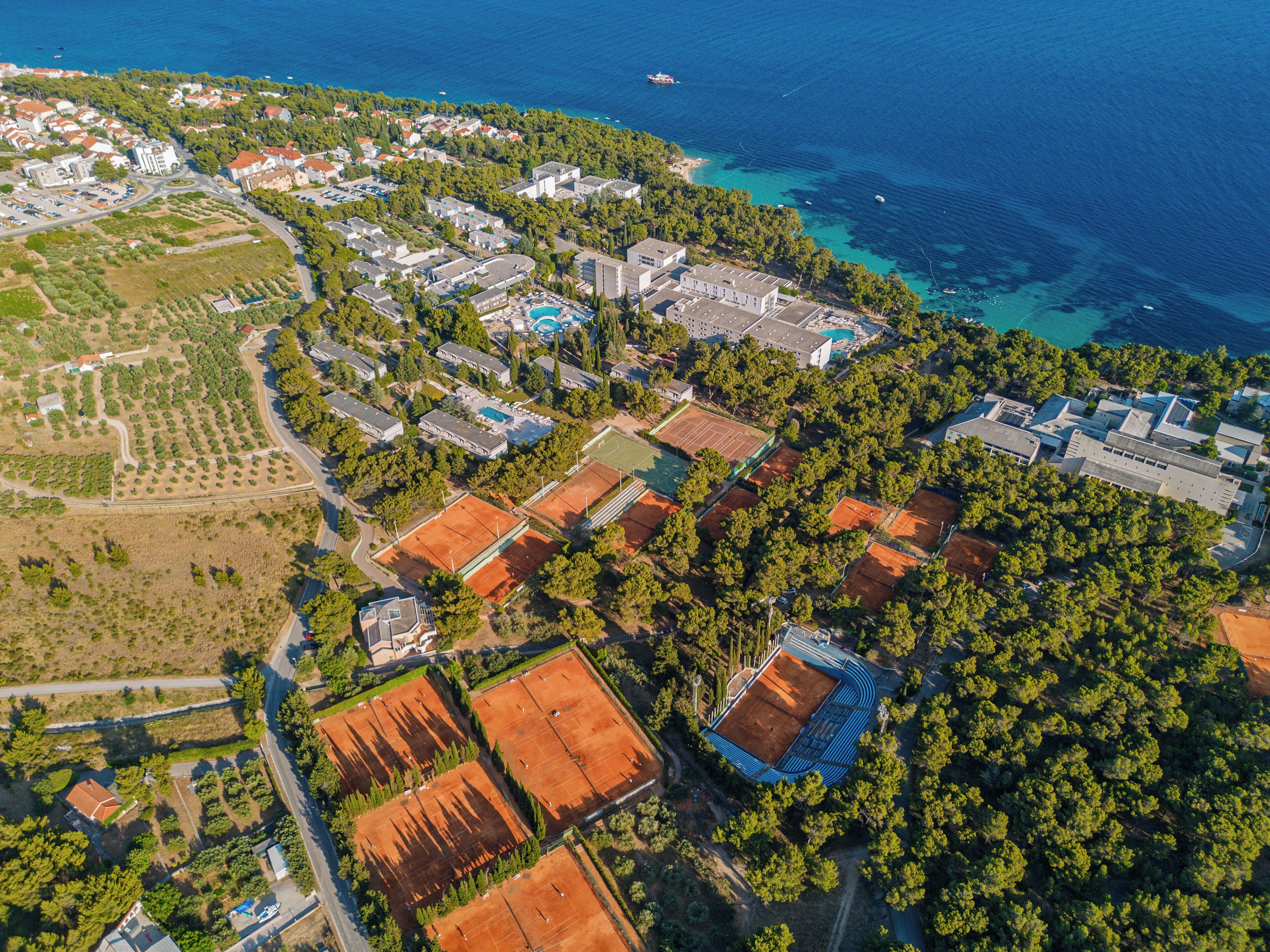 An aerial view of a coastal resort with multiple buildings, tennis courts, and a swimming pool.