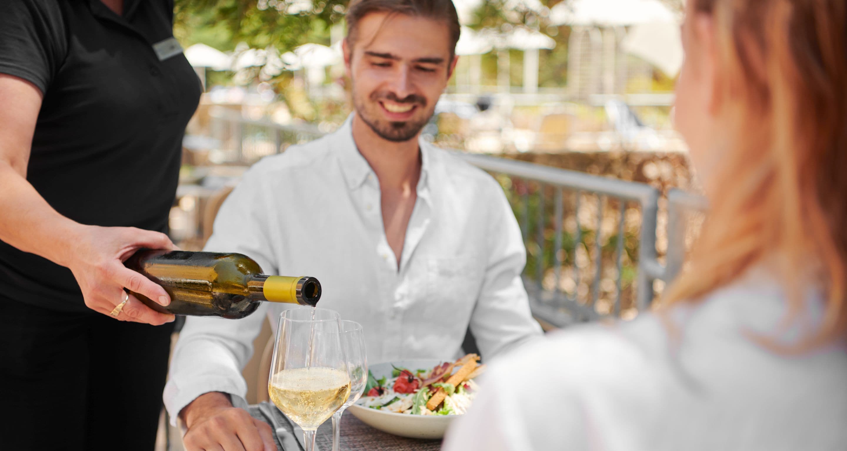 A person pouring wine into a glass at a table with a salad.