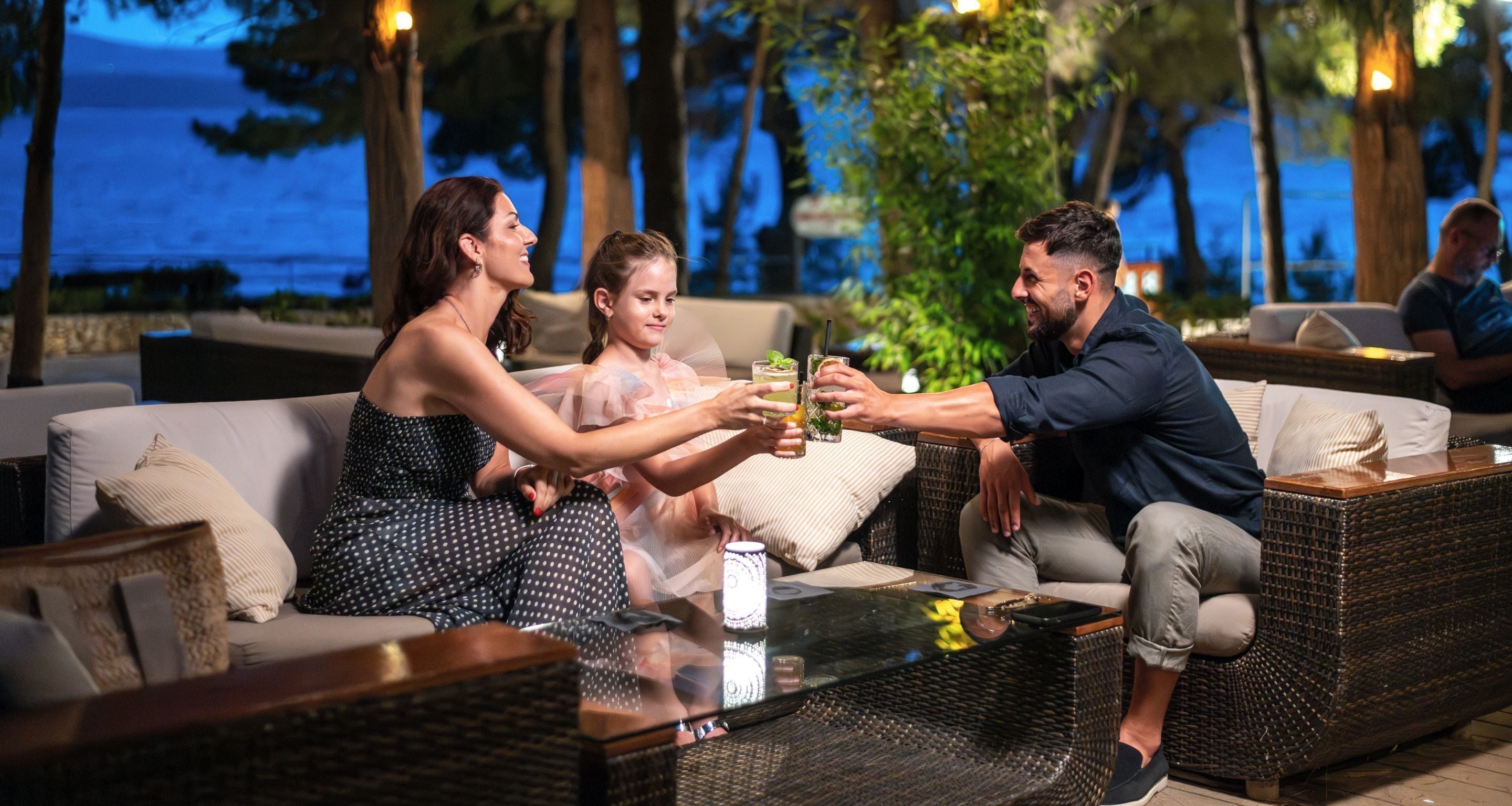 A group of people seated on a wicker sofa with a glass table in an outdoor setting with a view of the sea.