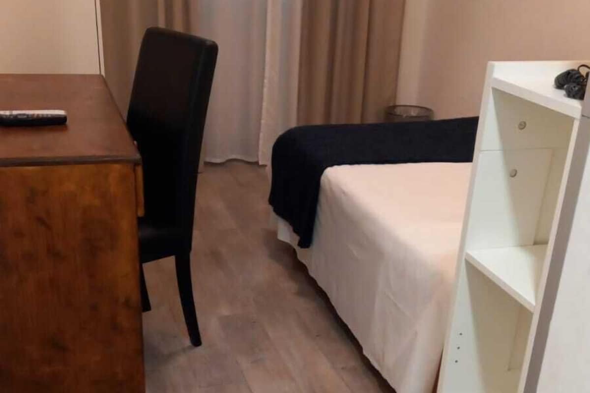 Una habitación de hotel con piso de madera, un escritorio con silla, una cama con cubrecama blanco y un armario blanco.