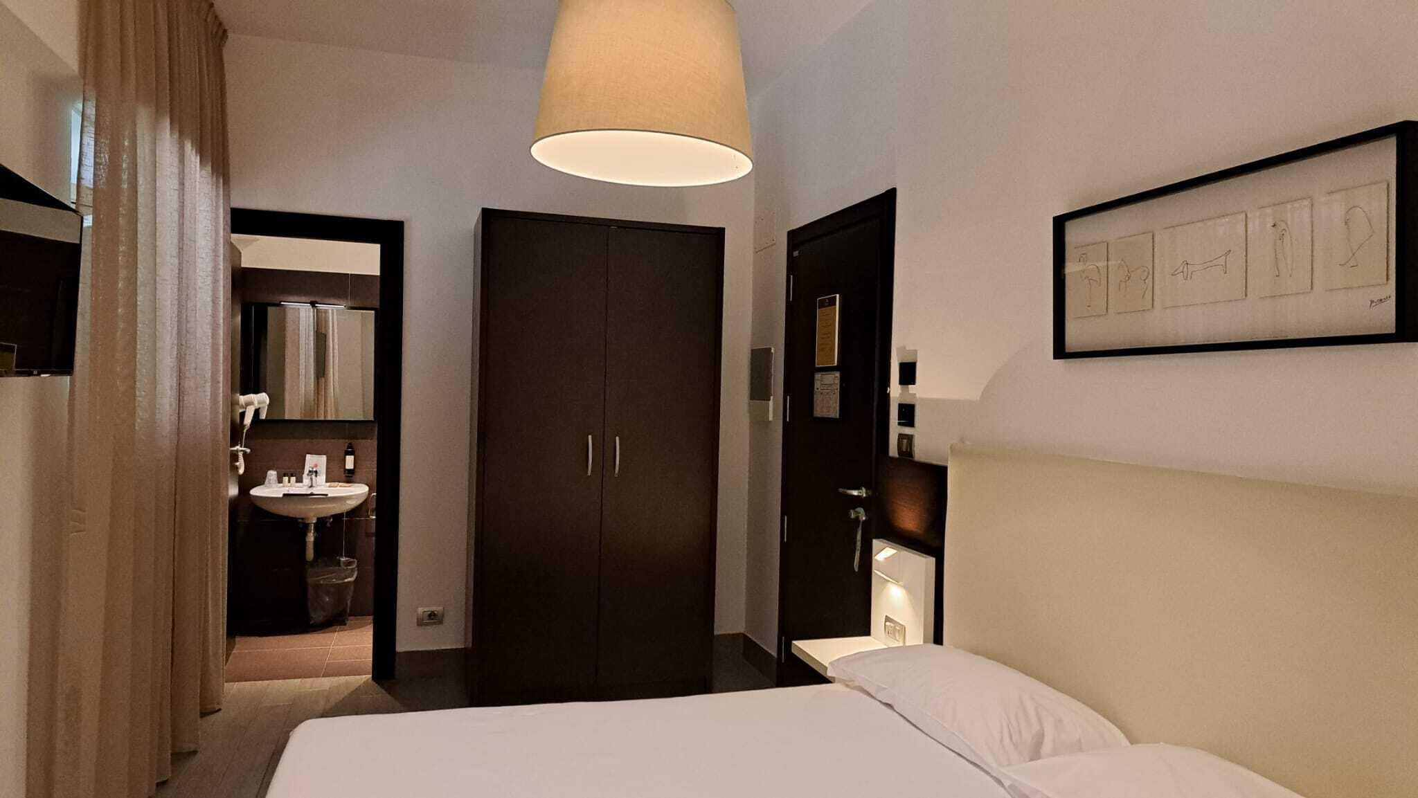 Hotelkamer met een bed, nachtkastje, groot raam, kledingkast en een zichtbare badkamer door een geopende deur.