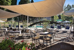 An outdoor patio area with tables and chairs, a pool in the background, and a covered seating area.