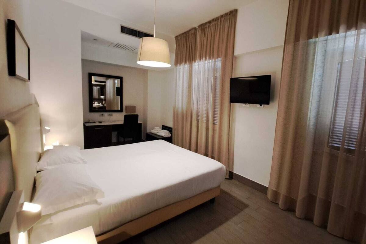 A hotel room with a bed, a desk, a chair, a mirror, a TV, and curtains.