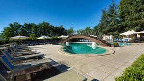 Un área de piscina con sillones de descanso y sombrillas, rodeada de árboles y un cielo azul despejado.