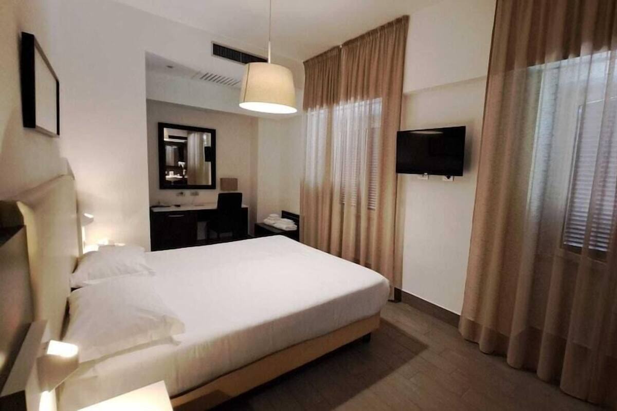 A hotel room with a bed, a desk, a chair, a mirror, a TV, and curtains.