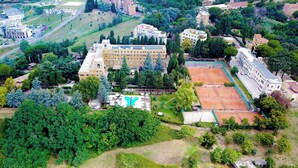 Vista aérea de un extenso complejo de edificios rodeado de vegetación, con varias canchas de tenis y una piscina.