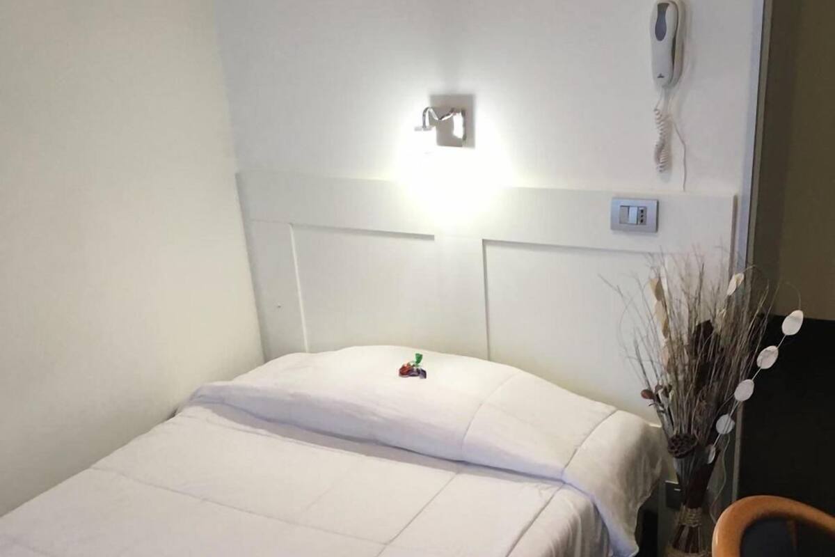 A hotel room with a bed, a bedside table, a telephone, and a vase with dried plants.
