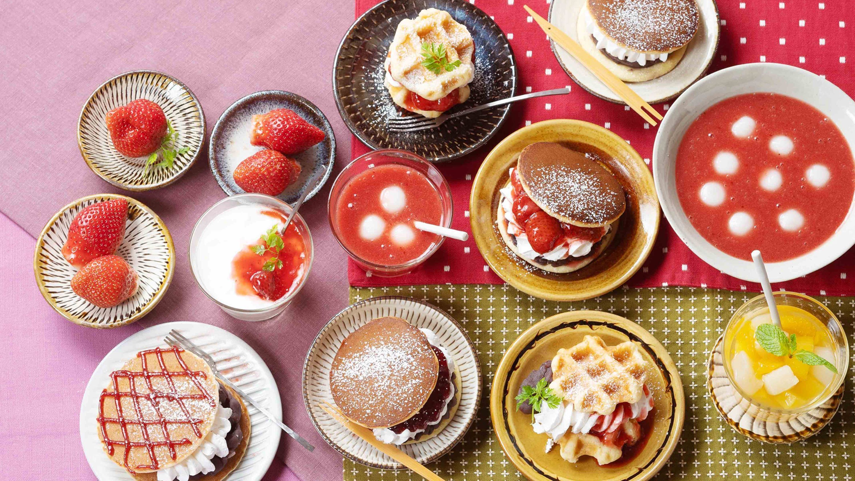 Une variété de desserts, notamment des gaufres, des crêpes et une soupe aux fraises, présentés sur une table recouverte d’une nappe à carreaux rouges et blancs.