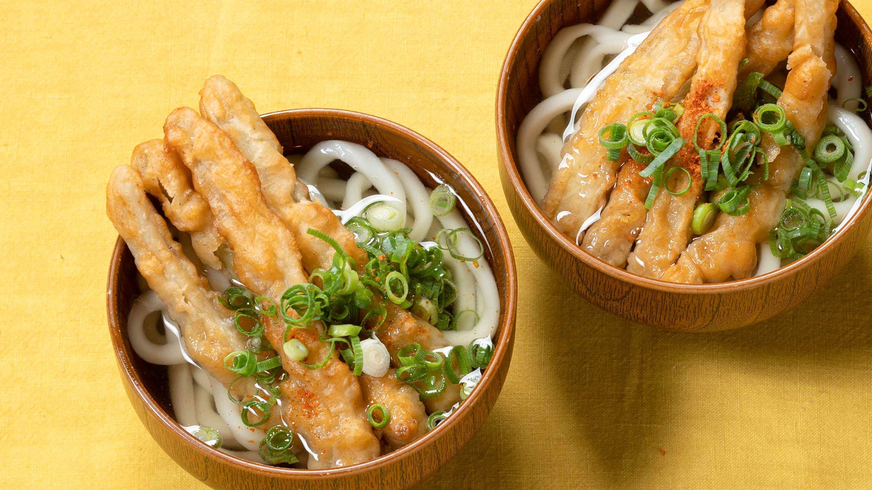 Two bowls of ramen with noodles, a protein, and garnishes on a yellow surface.