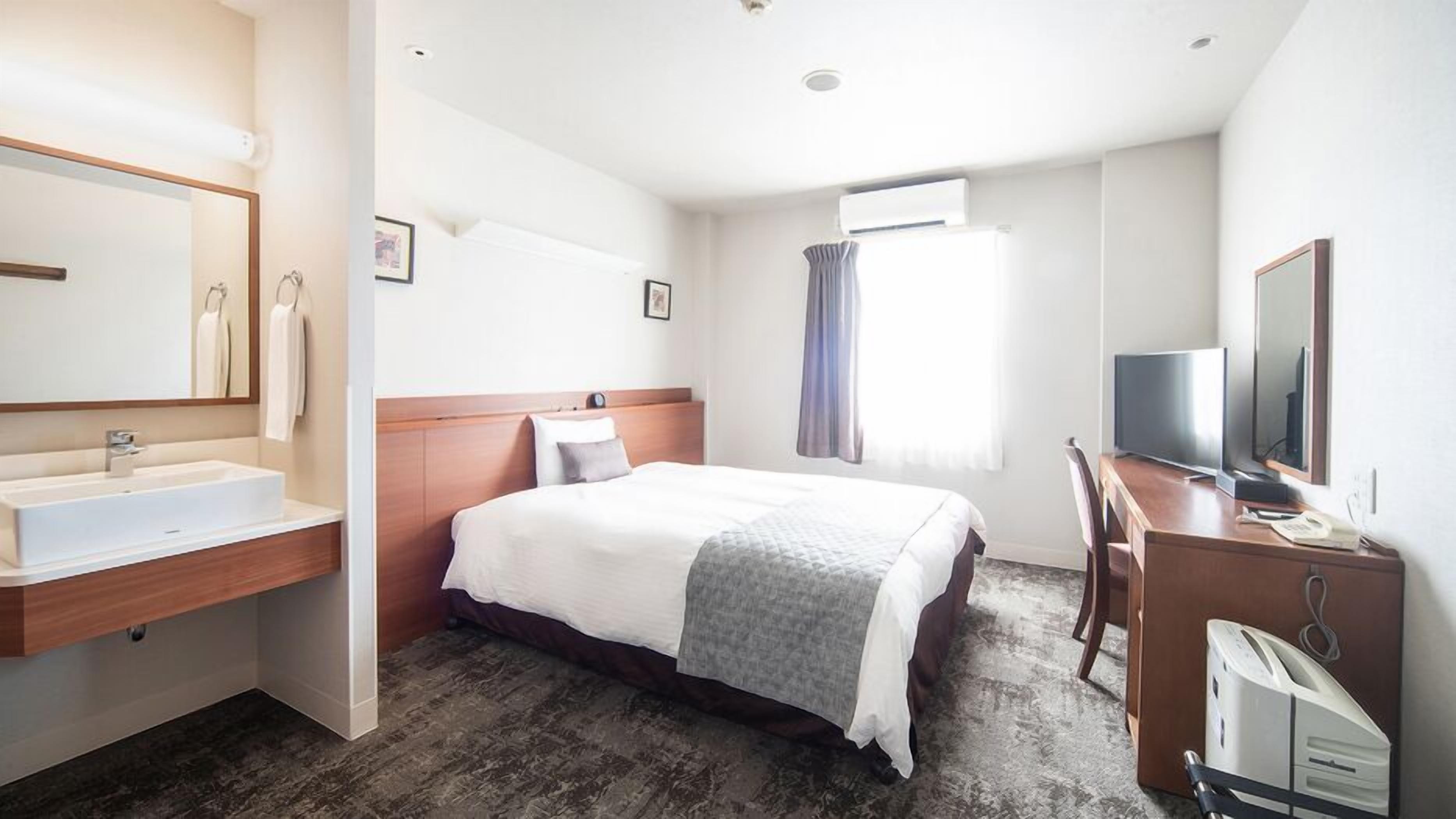 A hotel room with a bed, a desk with a TV, a bathroom with a sink, and a window with curtains.