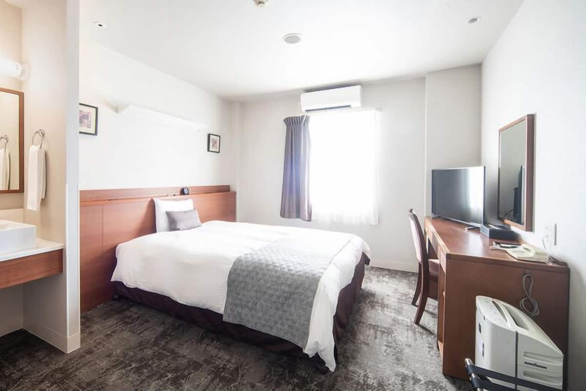 Ein Hotelzimmer mit Bett, Schreibtisch mit Fernseher, Badezimmer mit Waschbecken und Fenster mit Vorhängen.