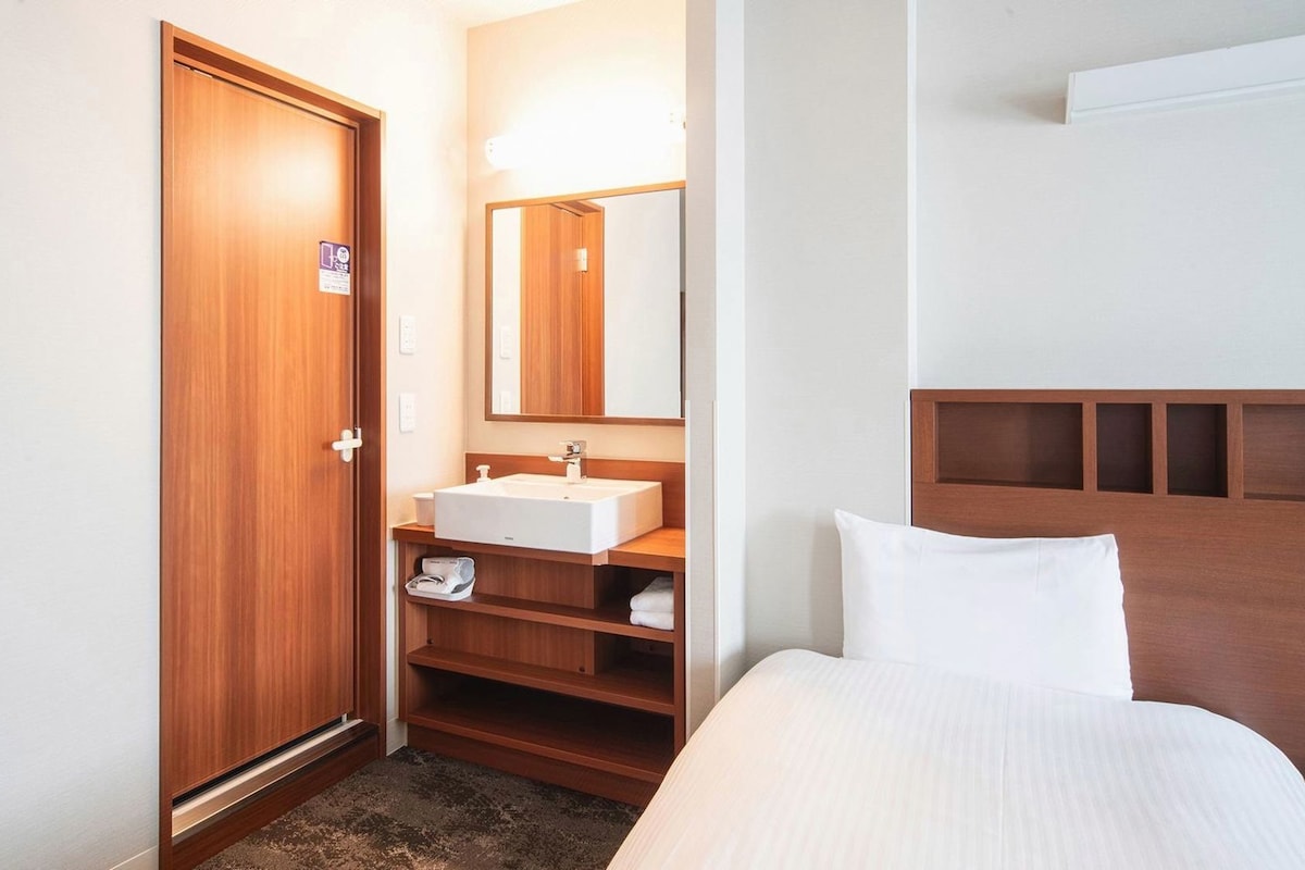 Ein Hotelzimmer mit Holztür, Waschbecken in Weiß, Spiegel, Bett mit weißer Bettwäsche und einem Kopfteil mit Stauraum.