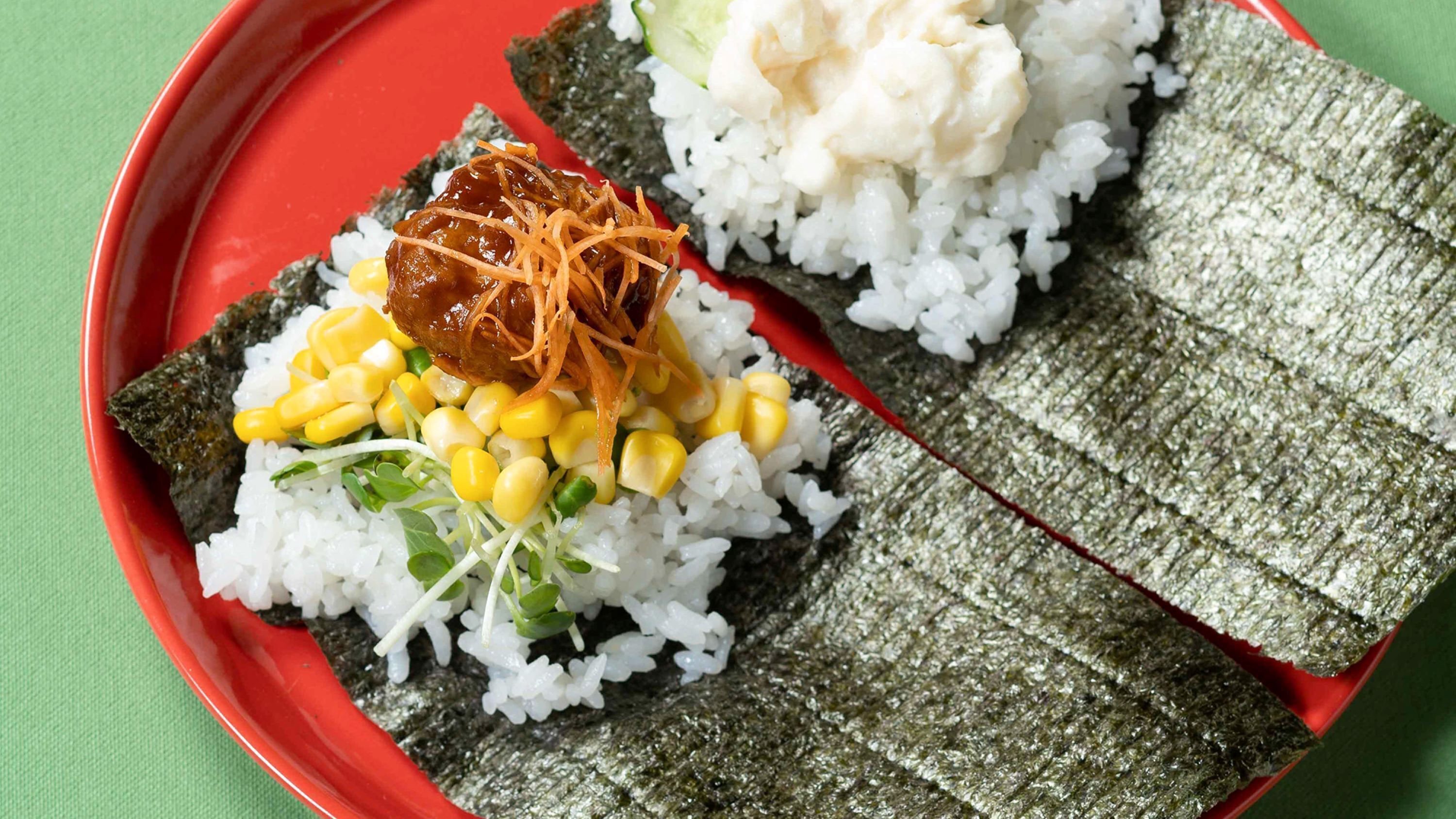 A red plate with two servings of rice wrapped in seaweed, one with corn and shredded carrots, and the other with a white topping.