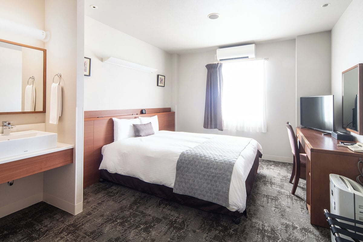 Ein Hotelzimmer mit Bett, Schreibtisch mit Fernseher, Spiegel, Badezimmer mit Waschbecken und Klimaanlage.