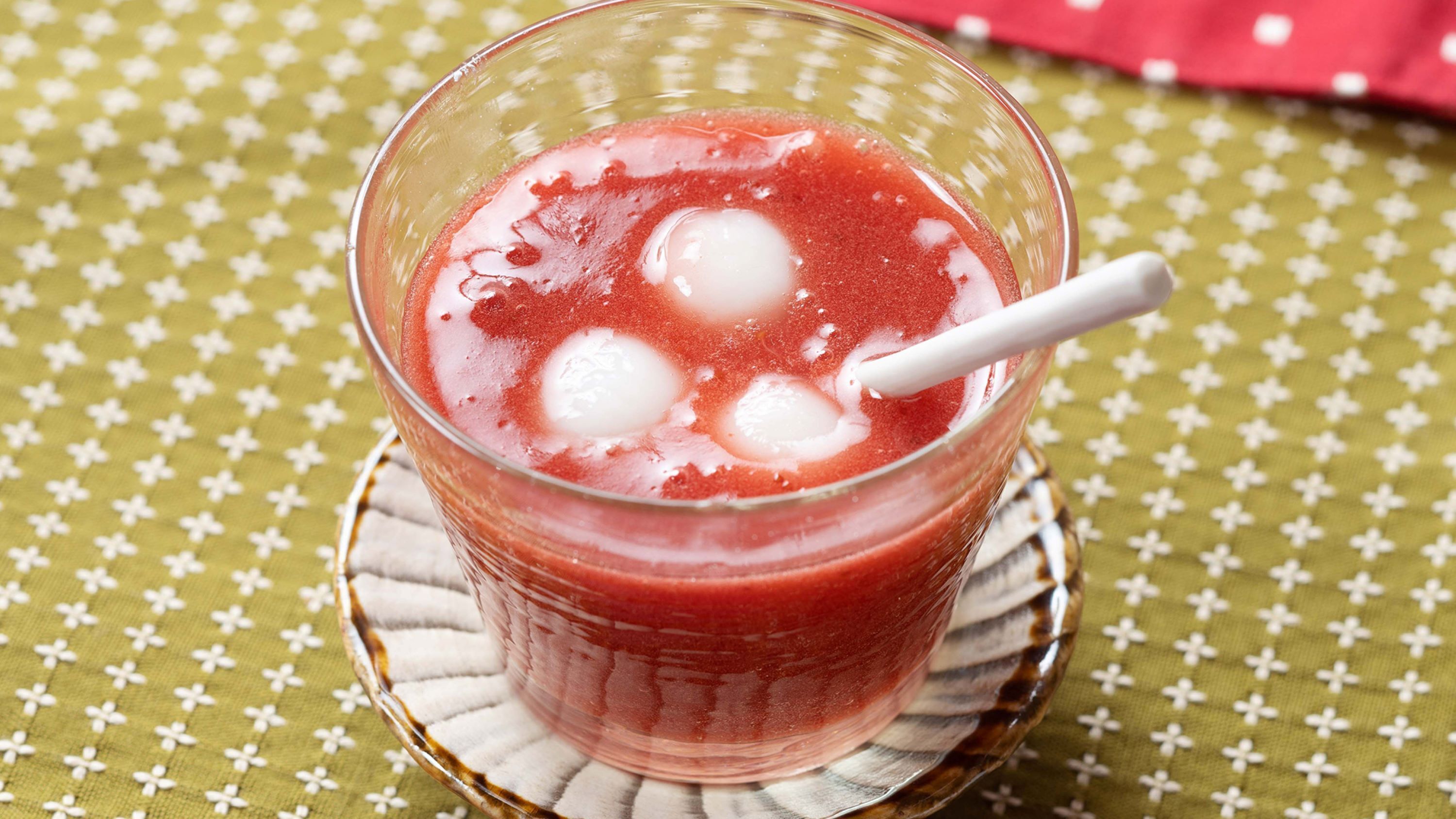 A glass of red drink with ice cubes on a patterned tablecloth.