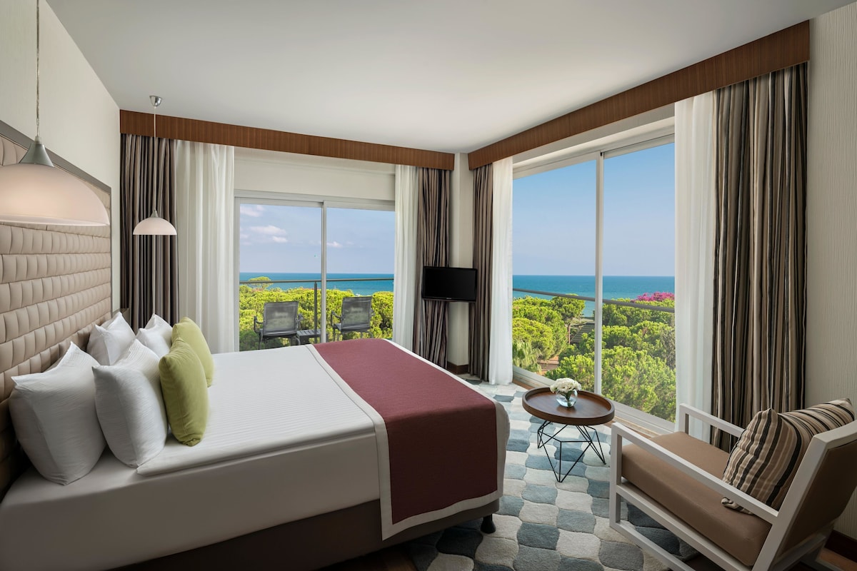 Habitació d'hotel amb un llit gran, un sofà, una taula petita, un televisor i un balcó amb vista al mar.