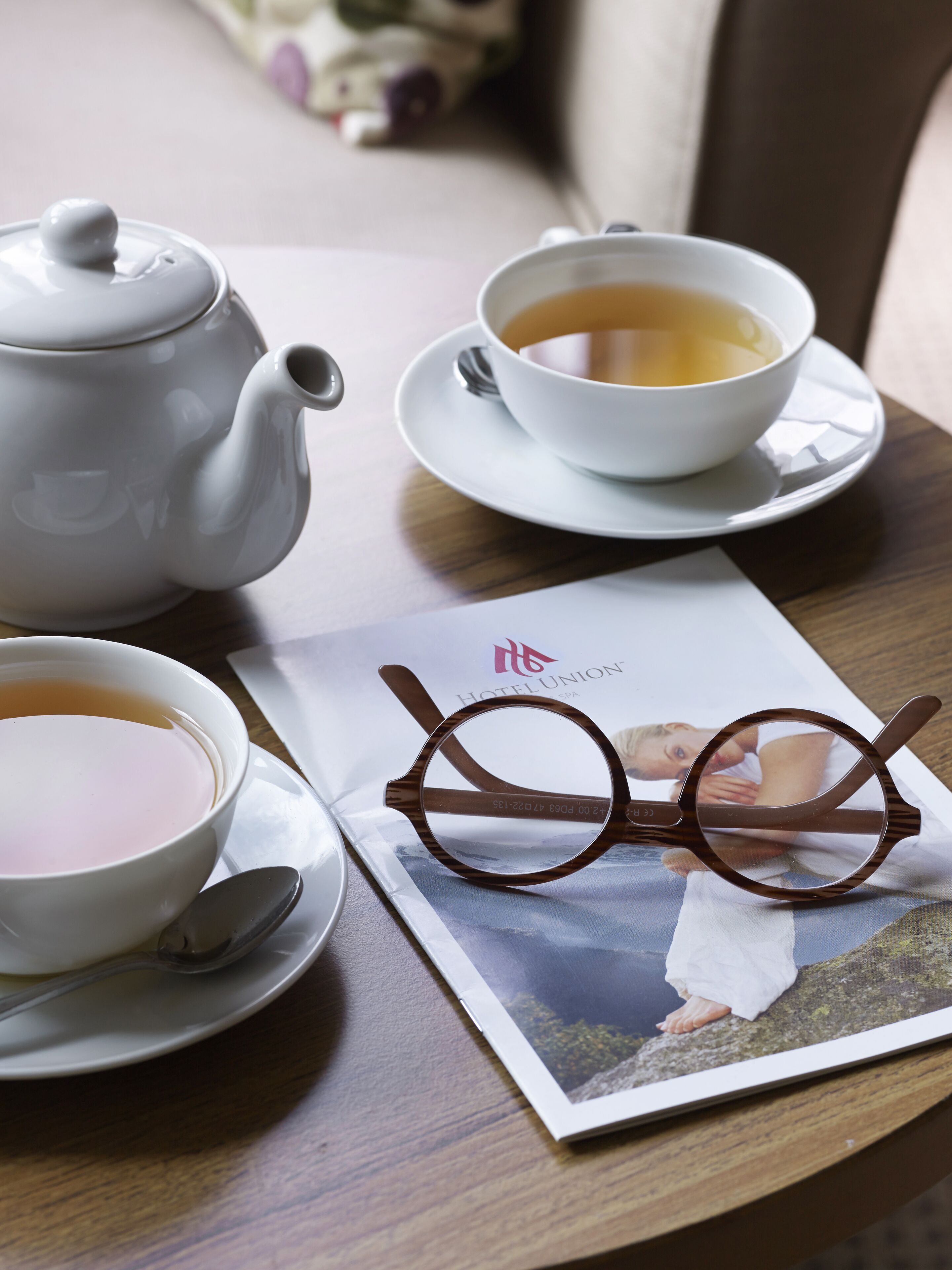A table with a teapot, a cup of tea, a spoon, and a pair of glasses on a magazine.