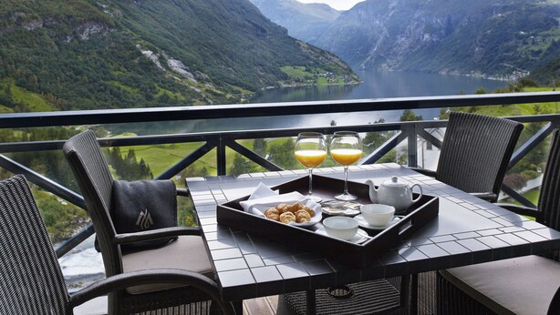 A dining table with breakfast items and glasses of orange juice, overlooking a mountainous landscape and lake.