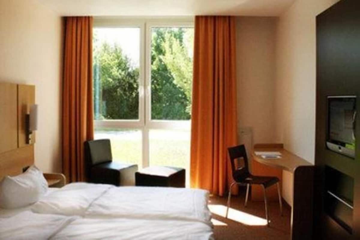 Una habitación de hotel con cama, escritorio, silla, televisor y ventana con cortinas.