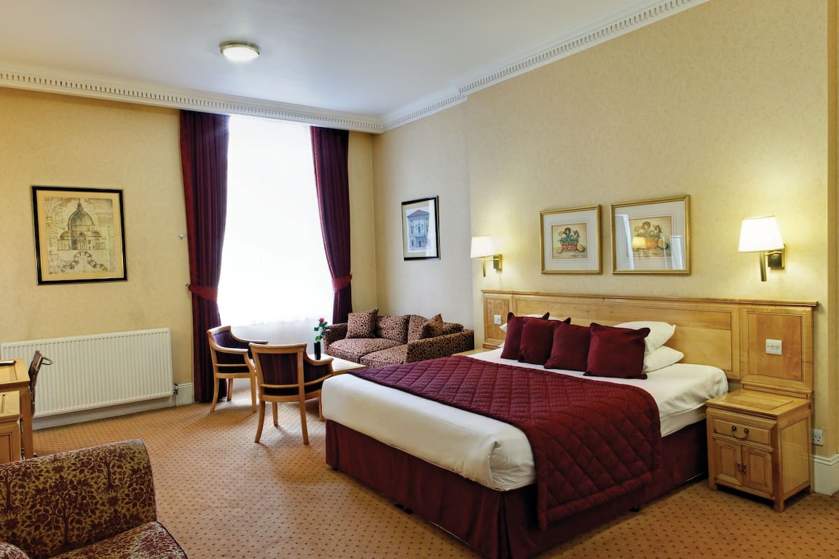 A hotel room with a large bed, a desk, a chair, and a sofa. There are two framed pictures on the wall and a window with curtains.