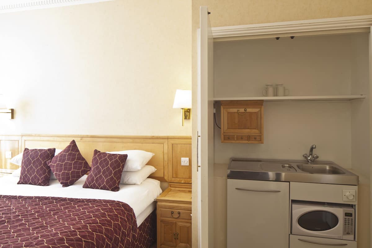 A hotel room with a bed, pillows, a nightstand, a lamp, a wooden cabinet, a microwave, and a sink.