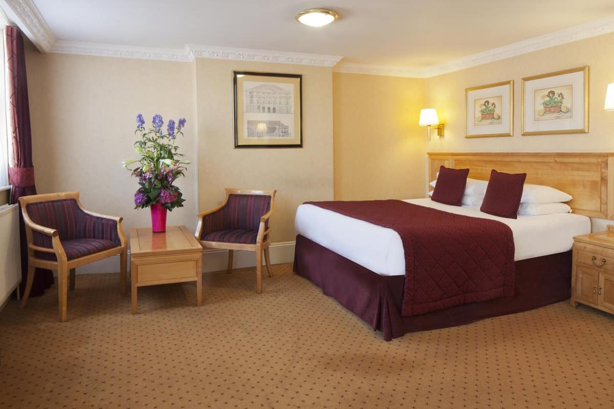 A hotel room with a large bed, two armchairs, a small table with a vase of flowers, a nightstand, and two wall-mounted lamps.