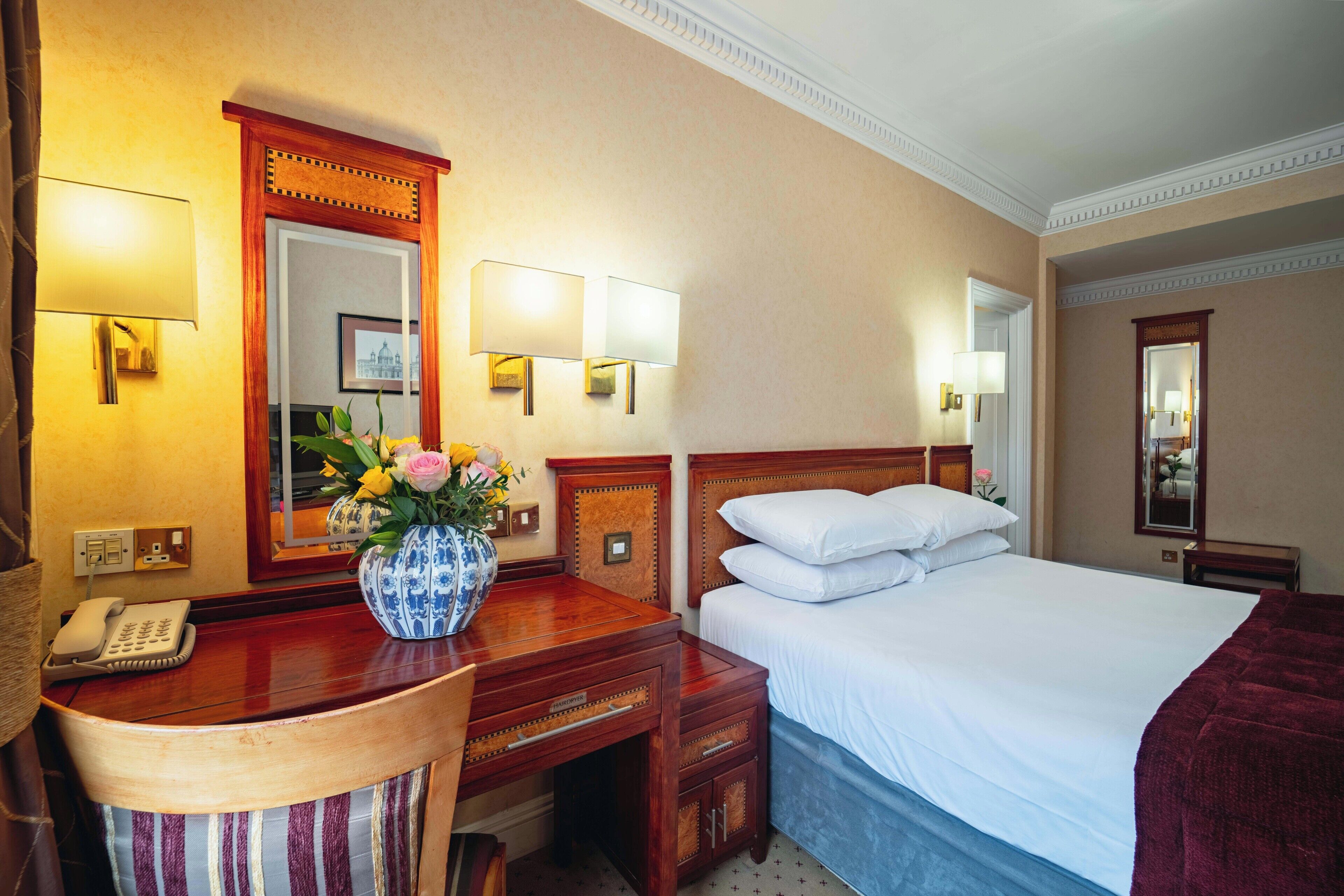 Une chambre d’hôtel avec un bureau en bois, un téléphone, un vase de fleurs, un lit avec du linge de maison blanc et deux lampes fixées au mur.