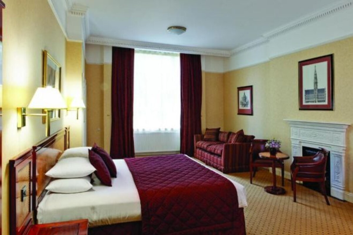 Ett hotellrum med en stor säng, ett skrivbord med en stol, en öppen spis och ett fönster med gardiner.