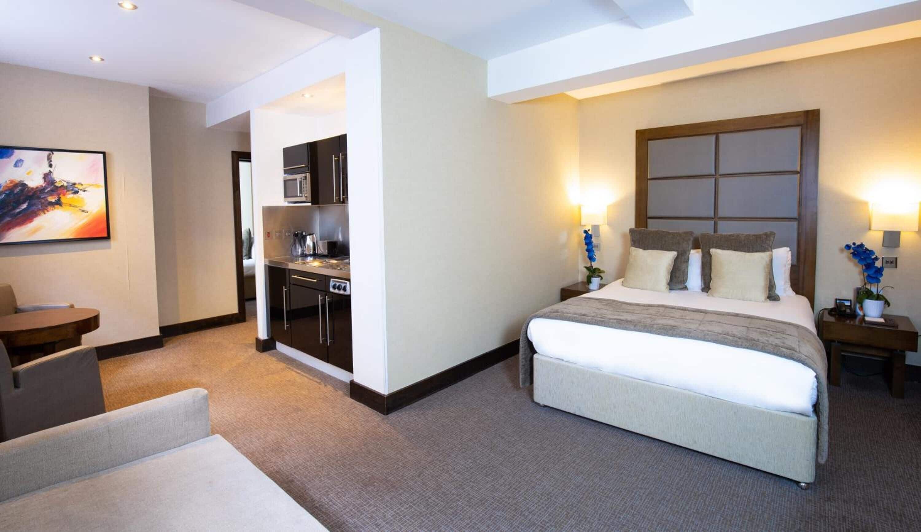 A hotel room with a large bed, a desk, a kitchenette, and a painting on the wall.