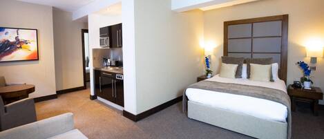 A hotel room with a large bed, a desk, a kitchenette, and a painting on the wall.