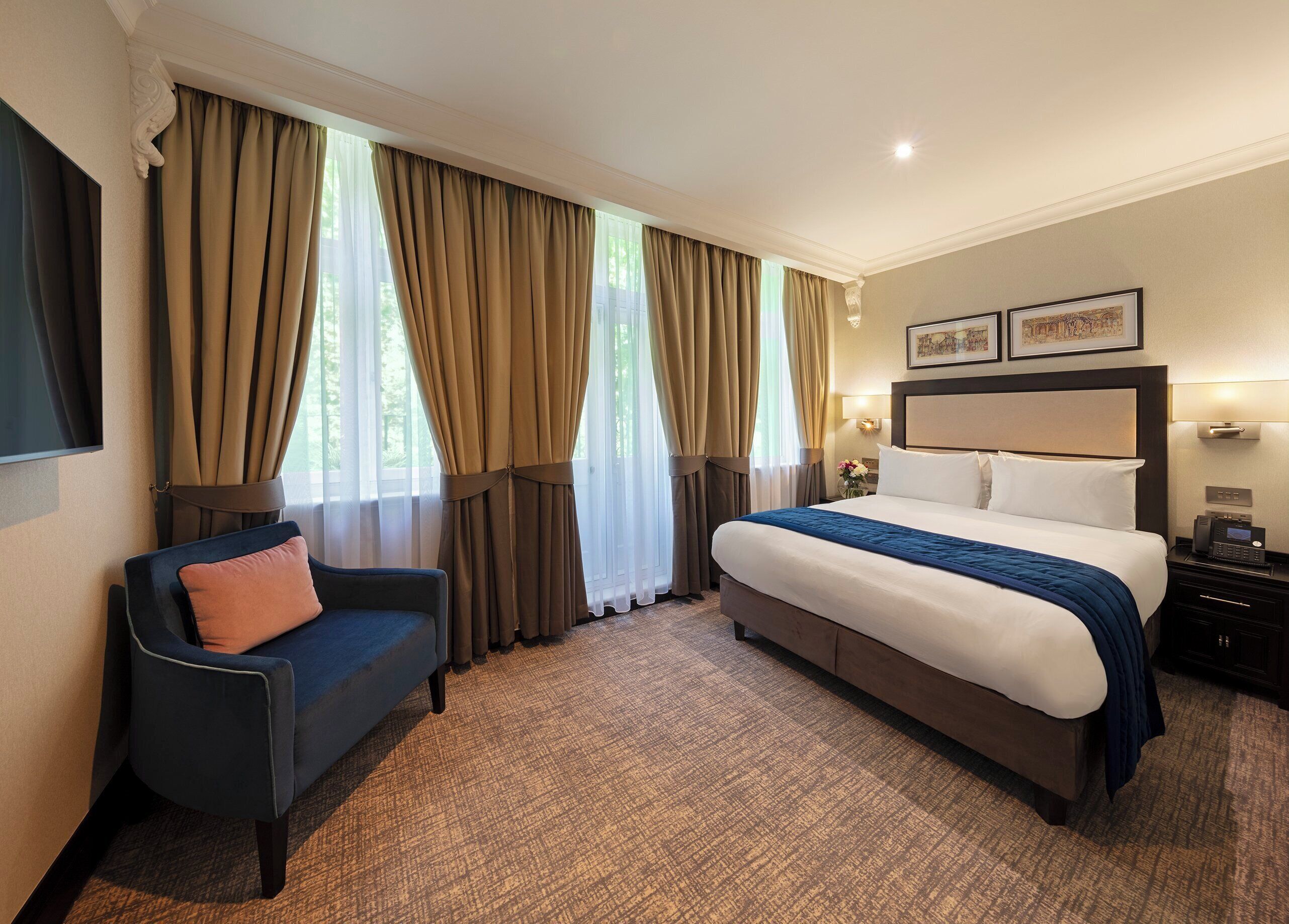 A hotel room with a large bed, a chair, a desk with a lamp, and curtains on the windows.