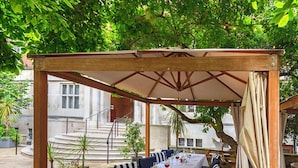 An outdoor dining area with a covered pergola, long tables set with white tablecloths, striped cushions, and wine glasses. - Wellington Hotel by Blue Orchid (London)