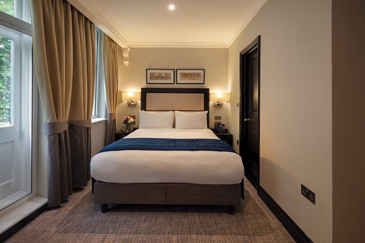 Habitación de hotel con una cama grande, dos mesitas de noche, un cabecero y una ventana con cortinas.