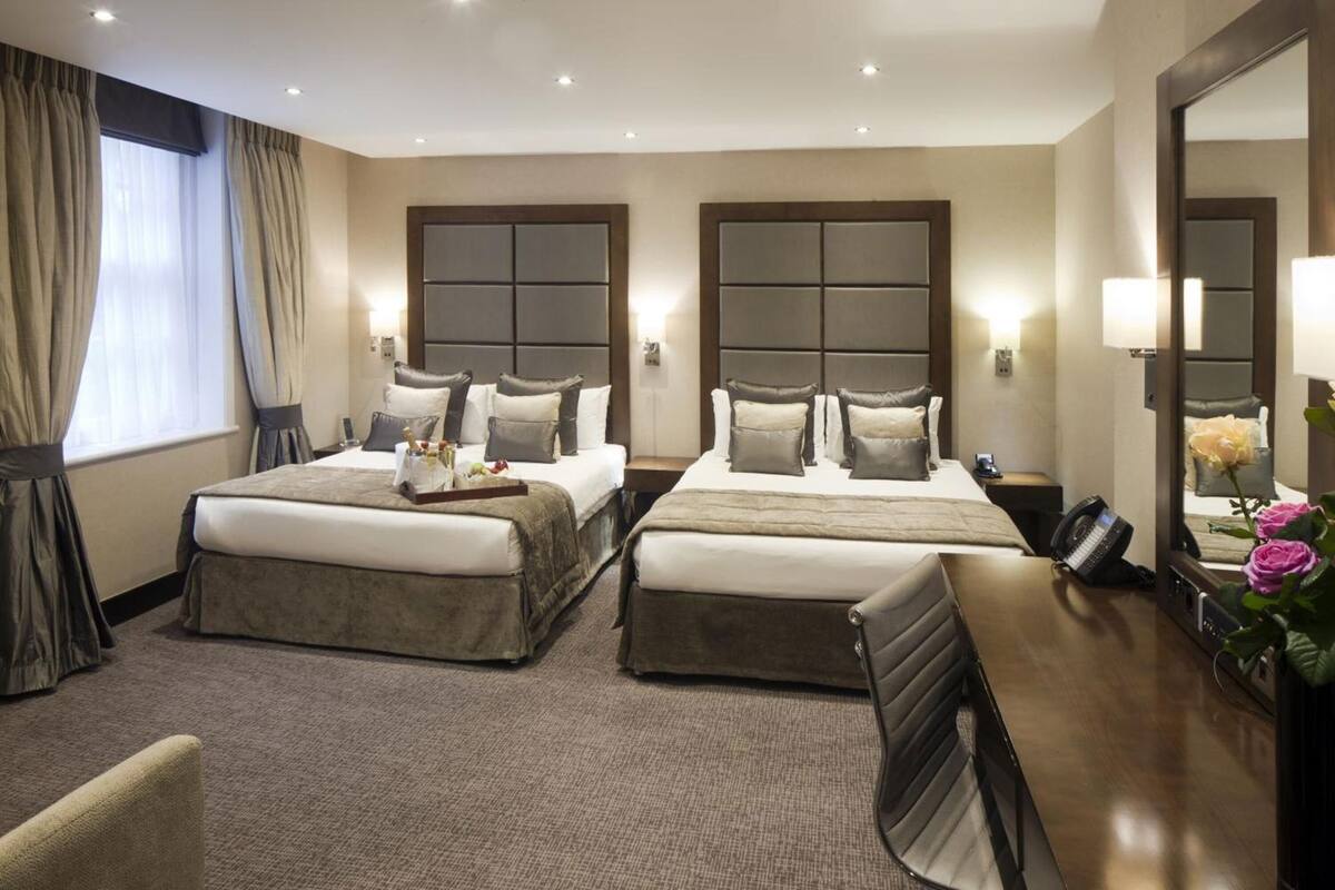 Habitación de hotel con dos camas, un espejo grande, un escritorio con teléfono y un florero con flores.