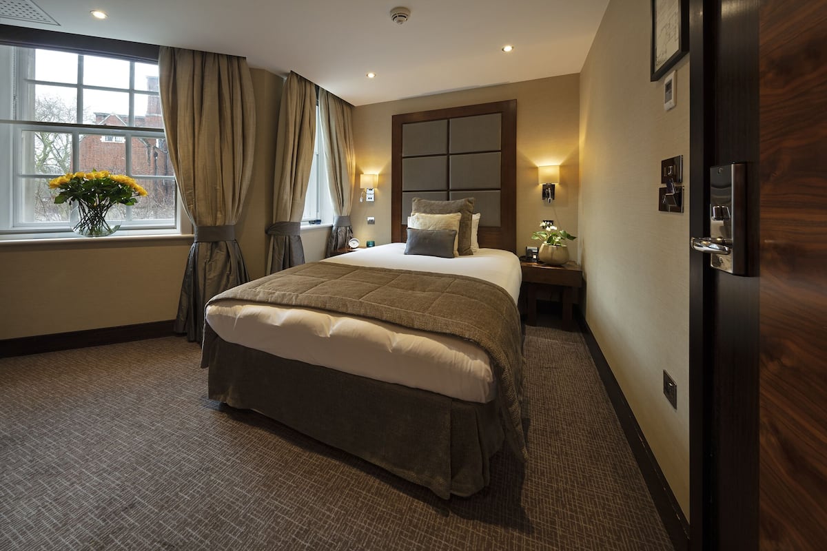 Habitación de hotel con una cama grande, una ventana con cortinas, una mesita de noche con lámpara y una lámpara de pared.