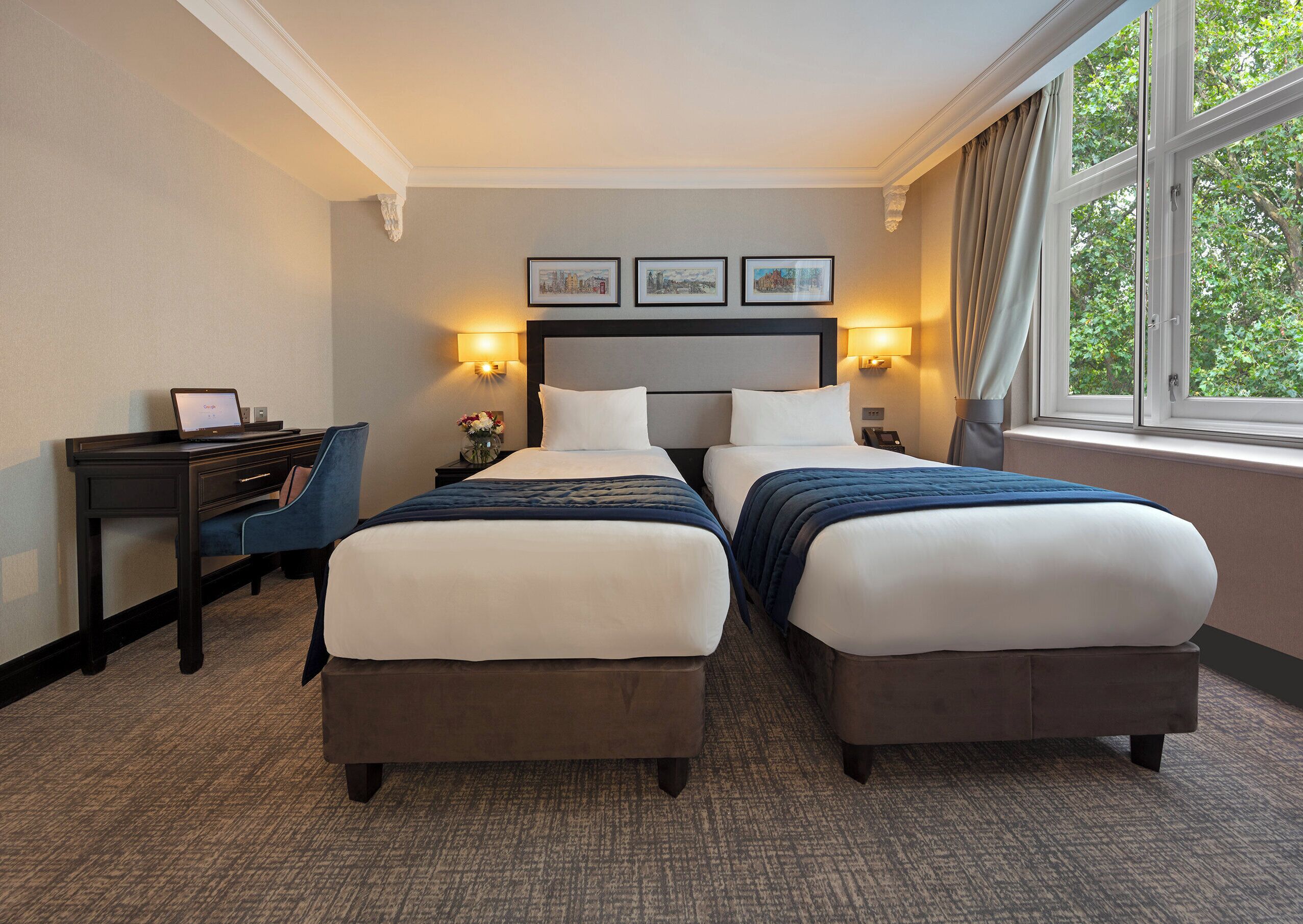 A hotel room with two beds, a desk, and a chair. There are wall-mounted lamps and a window with curtains.